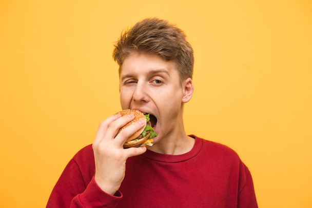 Yakın yukarı bir çocuk portre sarı bir arka planda bir burger yiyor ve kamera bakıyor, Genç adam bir burger ısırır. Fast-Food konsepti. - Fotoğraf, Görsel