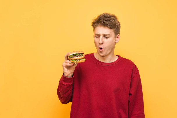 Funny young man holds a burger in his hands, looks at fast food with a surprised look, isolated on a yellow background. Hungry teenager with burger in his hands is on a yellow background. - Photo, Image