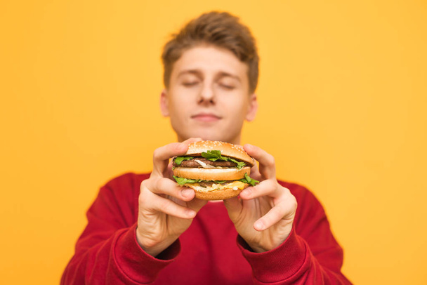 Handsome guy with his eyes closed shows a delicious large burger to the camera, holds in his hands, isolated on a yellow background.Focus on an appetizing burger in the hands of a young man. Fast food - Photo, Image