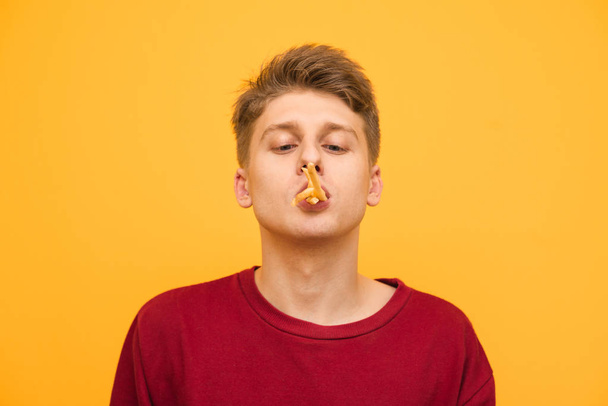Ritratto ravvicinato di un giovane divertente con le patatine fritte in bocca. allegro adolescente mangia un fast food, isolato su uno sfondo giallo. Copyspace
 - Foto, immagini