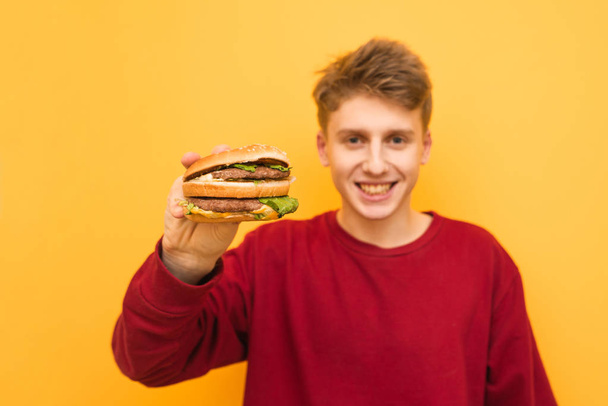 Happy Guy in Casual kleding heeft een smakelijke hamburger in zijn handen en toont in de camera, glimlachend, geïsoleerd op een gele achtergrond. Jonge man toont een grote smakelijke hamburger en kijkt naar de camera. - Foto, afbeelding