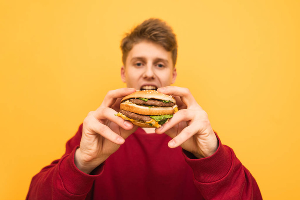 Hungry young man holds in the hands of a delicious big burger, looking at the camera on a yellow background. Focus on the sandwich in the hands of the guy, isolated. Background - Foto, afbeelding