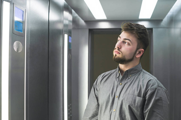 Retrato de um homem adulto barbudo vestindo uma camisa cinza fica em um elevador com um rosto sério e olha lado a lado no número no chão. Homem sério no elevador.
. - Foto, Imagem