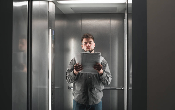 エレベーターの中に立っている書類を読む若い男性従業員の肖像画。集中したビジネスマンは、リフトのドキュメントをチェックしています. - 写真・画像