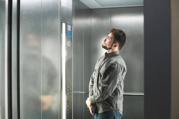 Man met een baard staat in de lift en kijkt naar het scherm met een vloer nummer, het dragen van een grijs shirt. Kantoormedewerker daalt in de lift. Man in de lift. - Foto, afbeelding