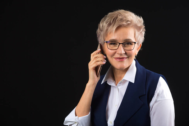 beautiful stylish aged woman in glasses in waistcoat on a black background calls on the phone - Fotó, kép