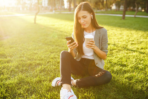 手に携帯電話とコーヒーを持つ緑の草の上に座って美しいスタイリッシュな女性の画像。彼女はワイヤレスヘッドフォンを通じて電話で話しています。サンセットライトライフスタイルコンセプト. - 写真・画像