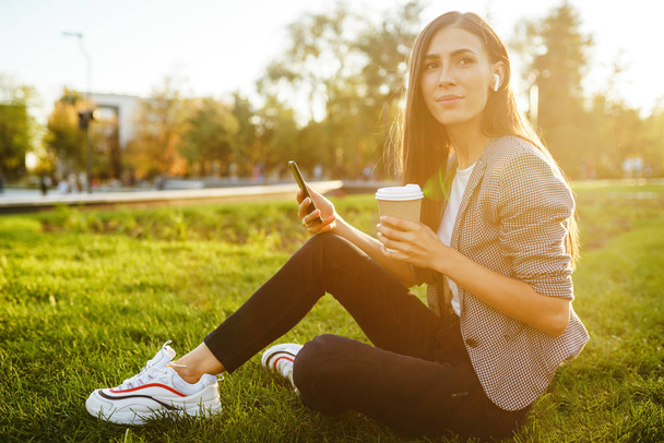 Image of beautiful stylish woman sitting on green grass with a phone in hand and coffee. She is talking on the phone through wireless headphones. Sunset light. Lifestyle concept. - 写真・画像