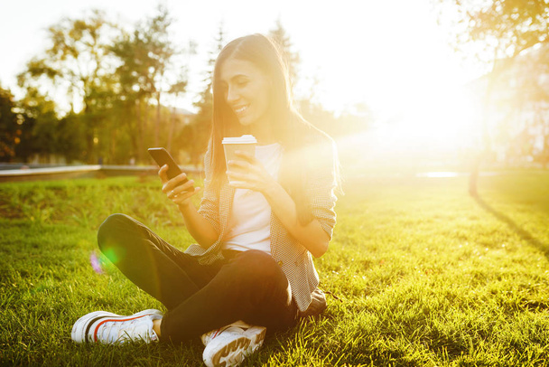 Image of beautiful stylish woman sitting on green grass with a phone in hand and coffee. She is talking on the phone through wireless headphones. Sunset light. Lifestyle concept. - Foto, Bild