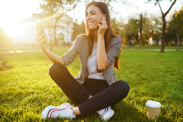 Image of beautiful stylish woman sitting on green grass with a phone in hand and coffee. She is talking on the phone through wireless headphones. Sunset light. Lifestyle concept. - Photo, image