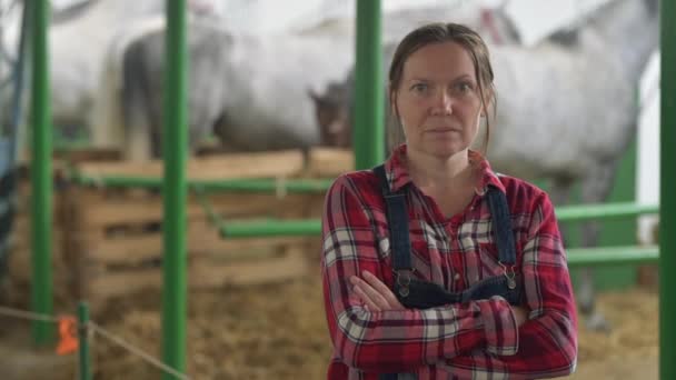 Portrait of female rancher at horse stable looking at camera. Adult woman wearing plaid shirt and jeans bib overalls as farm worker. - Video