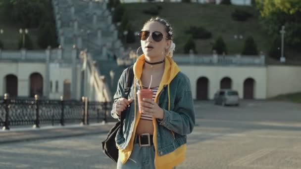 Gün batımında şehrin sokaklarında saçı örgülü genç bir kadın yürüyor. Güneş gözlüklü güzel kız şehir yürüyüşünde kahve içiyor. Orta boy. Yaşam tarzı kavramı - Video, Çekim