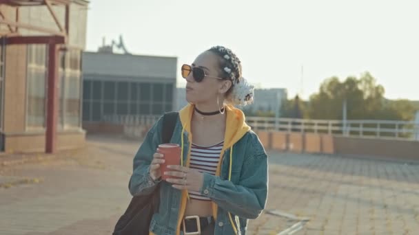 Hipster girl in jeans, hoodie with braids outdoors. Attractive young woman walking at sunny city in sunglasses with cup of tea or coffee. Medium shot - Video