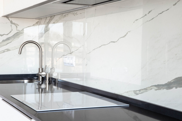 Close-up van witte glanzende keuken met zwarte Quartz countertop en marmeren tegel Backsplash. Ingebouwd verborgen kap en ondergemonteerde spoelbak.  - Foto, afbeelding