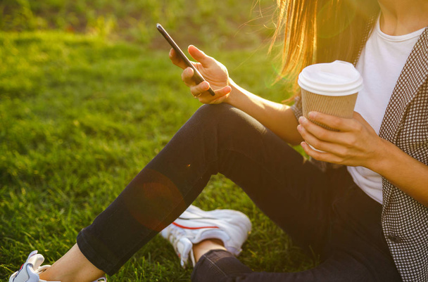 The girl holds the phone and coffee in hand. Image of beautiful stylish woman sitting on green grass with a phone in hand and coffee.  Sunset.  Summertime. - Фото, изображение