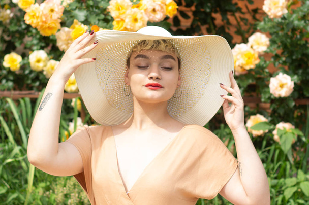 Uma bela jovem loira em um chapéu de abas largas expressa emoções em um jardim de rosas no fundo de um verão incrível. Brilhante sol e rosas amarelas. Retrato de moda ao ar livre glamour
. - Foto, Imagem
