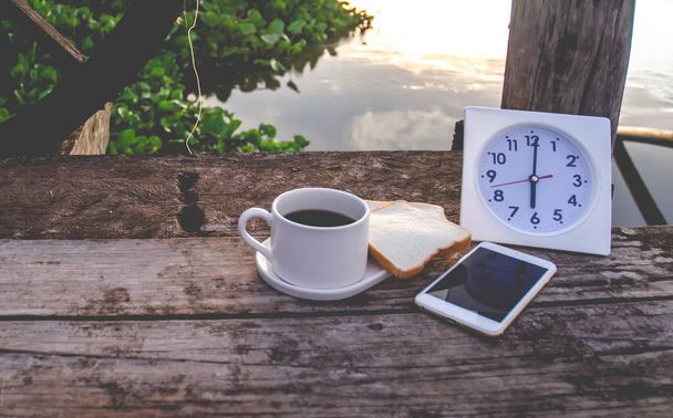 Kaffee Brot und weiße Uhr auf einem Holztisch am Morgen. Auf dem Fluss um 18: 00 Uhr gibt es eine weiße Uhr auf dem Holzboden. Smartphone auf den Tisch gelegt. Konzentrieren Sie sich nicht auf Objekte. Warmer Ton. - Foto, Bild