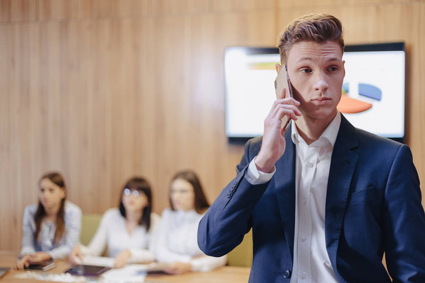 Stylish young businessman wearing a jacket and shirt on the background of a working office with people talking on a mobile phone - 写真・画像