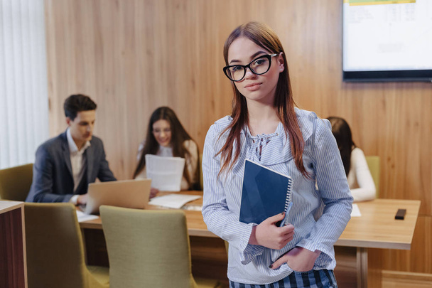 junge attraktive stilvolle Büroangestellte Mädchen in Brille mit einem Notizbuch in den Händen auf Hintergrund der Arbeitskollegen - Foto, Bild
