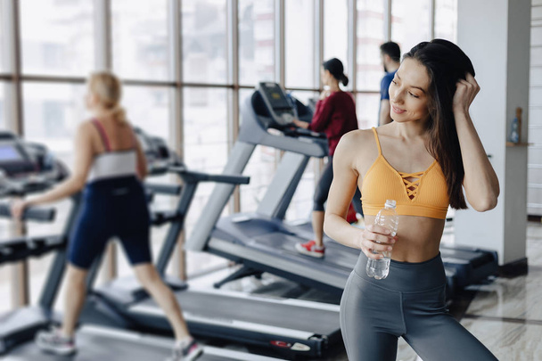 young attractive girl in the gym drinking water on the background of treadmills - Фото, изображение