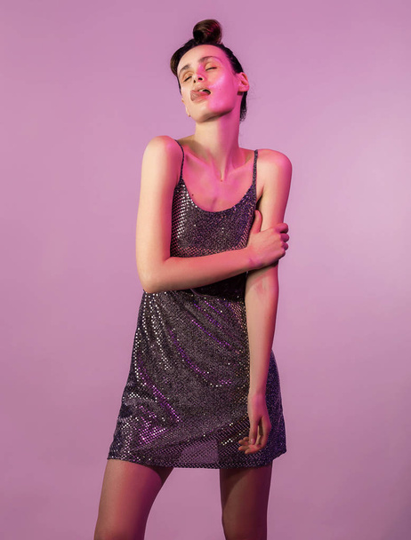 Fashion young girl put up a tongue in colorful bright neon lights, posing in studio. Girl in a brilliant dress hair tied in knot, gloss face in purple color light on pink background - Фото, изображение