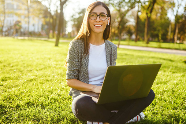 Image of beautiful stylish woman sitting on green grass with laptop  She is talking on the phone through wireless headphones. Sunset light. Lifestyle concept. - Photo, image