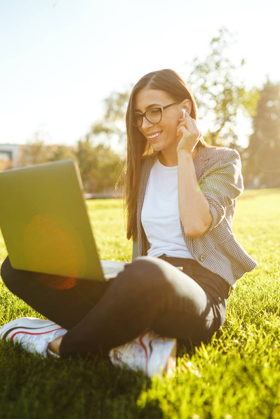 Image of beautiful stylish woman sitting on green grass with laptop  She is talking on the phone through wireless headphones. Sunset light. Lifestyle concept. - Foto, Imagem