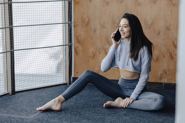 Mladá atraktivní fitness dívka, která sedí na podlaze u okna na pozadí dřevěné zdi, odpočívá ve třídách jóga a mluví po telefonu - Fotografie, Obrázek