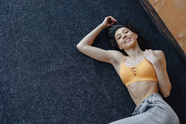 Young attractive fitness girl lying on the floor near the window on the background of a wooden wall, resting on yoga classes - 写真・画像