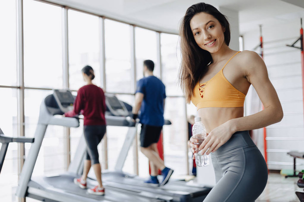 young attractive girl in the gym drinking water on the background of treadmills - Φωτογραφία, εικόνα