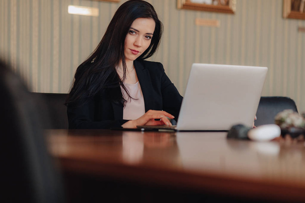 Mladá atraktivní emotivní dívka v šatech z obchodního stylu, která sedí u stolu na přenosném počítači a telefon v kanceláři nebo v hlediště - Fotografie, Obrázek