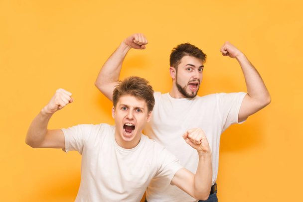 Joyful men in white T-shirts rejoice with their hands raised and look into the camera and stand on a yellow background. Emotional friends have won, happy on the yellow background. - Foto, immagini