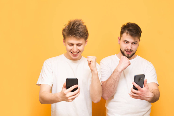 Muotokuva kahdesta onnellisesta miesystävästä, jotka seisovat keltaisella taustalla älypuhelimet käsissään ja iloitsevat. Kaksi ystävää käyttää valkoista t-paitaa, pitää älypuhelimet käsissään ja ovat onnellisia
. - Valokuva, kuva