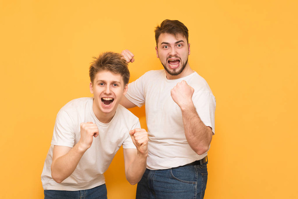 Portrait de deux hommes vainqueurs, se réjouissant les mains levées, regardant dans la caméra, portant un T-shirt blanc, sur un fond jaune. Deux amis heureux se réjouissent
. - Photo, image