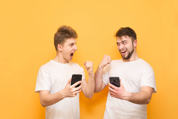 Δύο χαρούμενοι άνδρες κρατούν τα smartphones στα χέρια τους και κοιτάτε το ένα το άλλο, απομονωμένα σε κίτρινο φόντο. Οι χαρούμενοι άνθρωποι σε ένα κίτρινο φόντο χαίρονται με smartphones στα χέρια τους. - Φωτογραφία, εικόνα