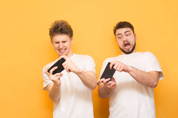 Twee verraste jonge mannen op een gele achtergrond dragen een wit T-shirt en spelen spelletjes op een smartphone. Emotionele vrienden spelen mobiele games, geïsoleerd op een gele achtergrond - Foto, afbeelding