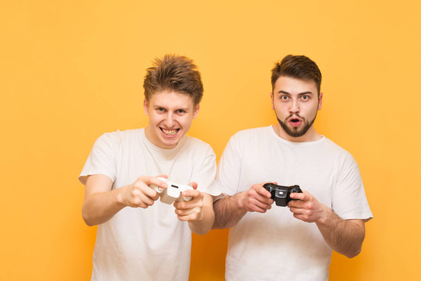 Dwóch emocjonalnych graczy z gamepadem w ręku, grając w gry wideo koncentruje się na żółtym tle patrząc w aparacie. Dwóch znajomych grać w gry na konsoli, z joysticki w ich rękach, izolowane - Zdjęcie, obraz