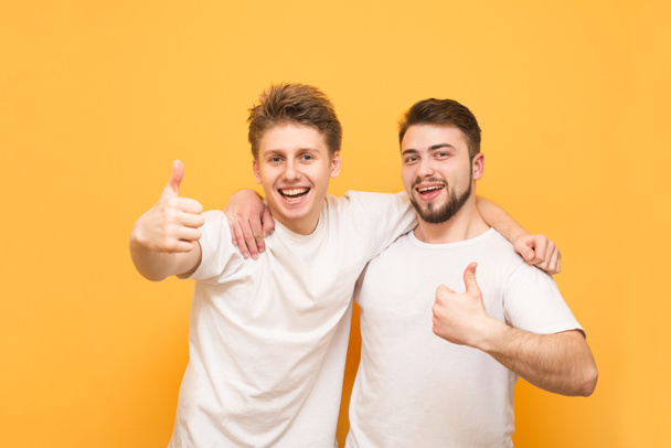 Portrait of a two happy young men showing thumbs up isolated over yellow background. Two happy friends in white t-shirts hugged by men, looking at the camera, smiling and showing a thumbs up - Photo, Image
