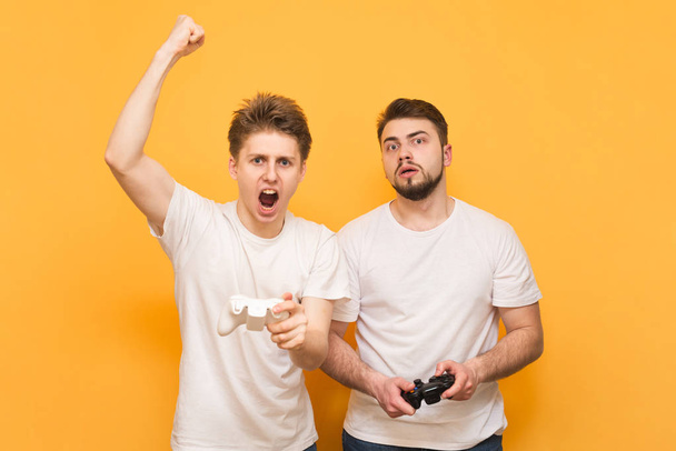 Two emotional friend with joysticks in their hands play video games, isolated on a yellow background. Winner and loser are playing video games, happy gamer and sad are on a yellow background. - Photo, Image