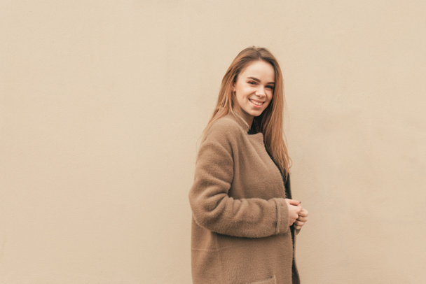 Attractive happy girl in a coat stands on the background of a beige wall, looks at the camera and smiles. Isolated. Copyspace - Photo, Image