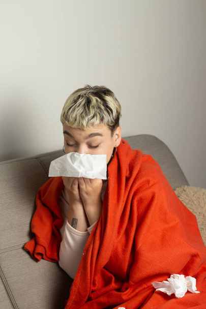 Unhealthy girl sitting on the couch, wrapped in a warm orange blanket, holding a napkin, sneezing and blowing her nose. Girl looks with chronic allergic rhinitis or cold difficult to breathe concept. - Photo, Image