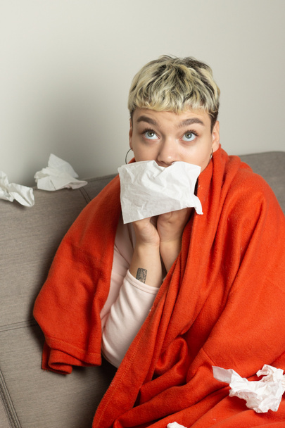 Unhealthy girl sitting on the couch, wrapped in a warm orange blanket, holding a napkin, sneezing and blowing her nose. Girl looks with chronic allergic rhinitis or cold difficult to breathe concept. - Photo, Image