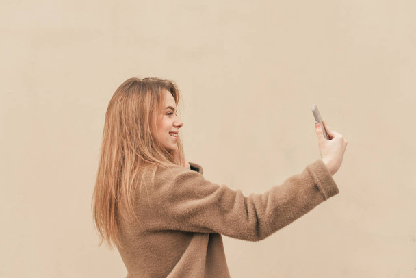 Sweet girl in a stylish spring dress, wearing a coat, standing on the background of a beige wall and takes selfie on a smartphone. Fashionable girl takes her photo on the street, background beige. - Фото, изображение