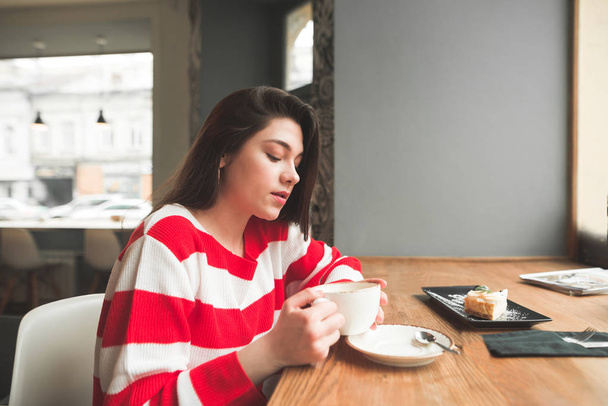 Houkutteleva brunette kuluvat rento vaatteita istuu kahvilassa ikkunan lähellä kahvia ja jälkiruokaa. Kaunis tyttö juo kahvia kahvilassa
. - Valokuva, kuva