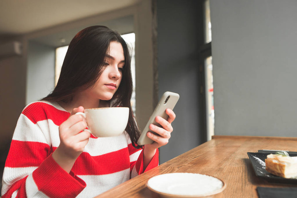 Young girl dressed in casual clothing sits in a cafe with a cup of coffee in her hands and uses the internet on a smartphone. Student sitting at a cafe table and looking at a smartphone screen. - Fotoğraf, Görsel