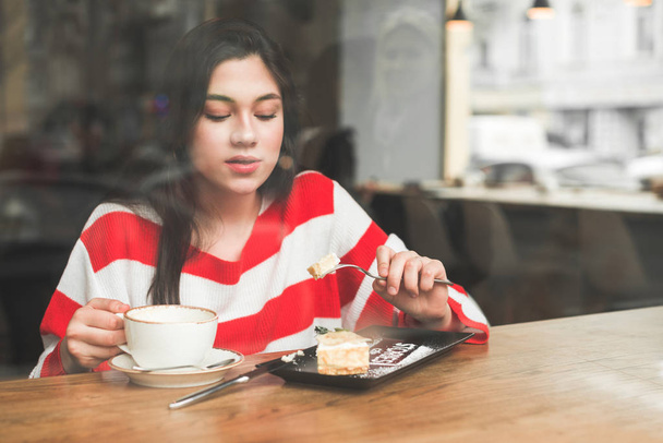 La chica se sienta en un acogedor café a la mesa cerca de la ventana, comiendo un postre apetitoso y sosteniendo una taza de café. Chica atractiva comiendo pastel en un restaurante y tomando café, mirando un postre
 - Foto, imagen