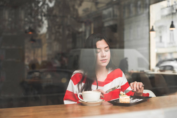 Sweet girl in casual clothes is having a rest in the cafe with a cup of coffee and a dessert on a day off. Portrait of a girl in a cafe through the restaurant window, looking at a plate with a dessert - Foto, Bild