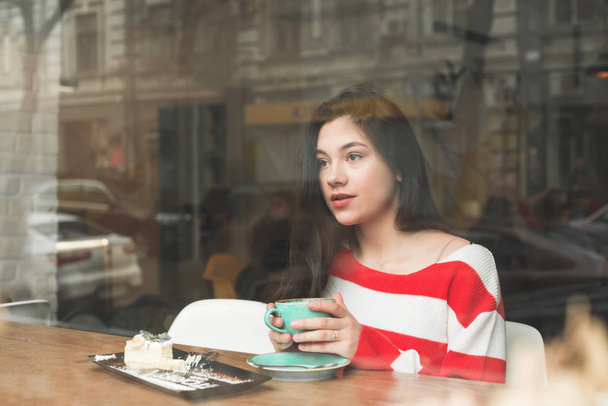 Красива дівчина проводить час в кафе, сидячи за столом з плитою з десертом і чашкою кави в руках, з нетерпінням задумливо в бік. Брюнетка страви в кафе. - Фото, зображення