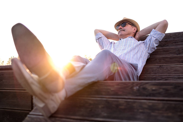 Viejo hombre hispano se sienta en el banco, sonriendo, disfrutando de un día soleado de verano
. - Foto, Imagen