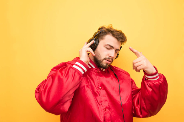 Ein bärtiger Mann in roter Jacke hört über Kopfhörer Musik und tanzt auf gelbem Hintergrund. Musikliebhaber hört Musik auf dem Hintergrund der gelben Wand und genießt. - Foto, Bild